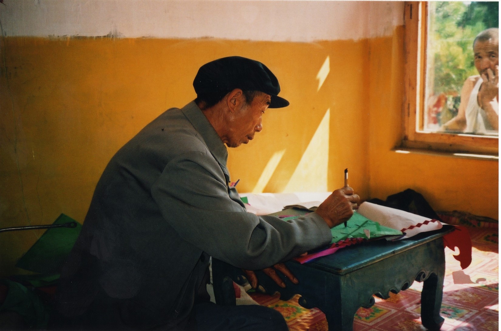 An image of a Chinese man wearing a cloth cap and pale blue jacket, sat at a desk writing. Another man is peering in through the window in front.